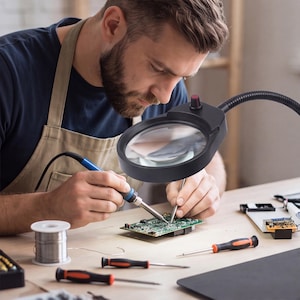 May include: A black magnifying lamp with a flexible neck and a top-mounted power switch, positioned over a green circuit board. The lamp features a circular lens frame and is used for detailed electronic repair work alongside a soldering iron.