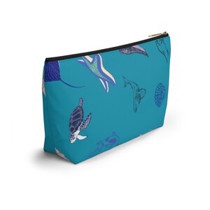 May include: A teal zippered pouch featuring a repeating pattern of various marine life illustrations, including a sea turtle, a stingray, a whale, a starfish, and a seashell, rendered in shades of blue, white, and purple against a solid teal background.