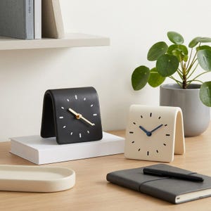 May include: Two arched tabletop clocks with a smooth finish. One clock is matte black with white hour markers and gold hands. The other clock is cream-colored with black hour markers and dark blue hands. Both clocks have a curved, folded shape.