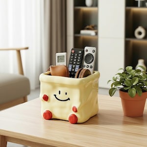 May include: A pale yellow ceramic storage container with a crinkled texture, featuring a black smiling face graphic and four red circular feet. The container holds a small brown wallet, a digital thermometer, and two black and white remote controls.