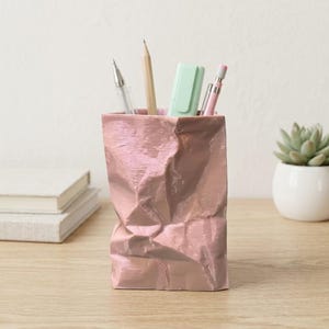 Könnte beinhalten: Ein Stiftehalter in metallischem Roségold, der wie eine zerknitterte Papiertüte geformt ist. Der Behälter hat eine texturierte, zerknitterte Oberfläche und enthält verschiedene Schreibgeräte, darunter einen Bleistift, einen hellgrünen Textmarker und Stifte.