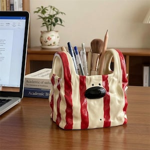 May include: A ceramic desk organiser shaped like a crumpled shopping bag with vertical red and cream stripes. It features a glossy black oval nose detail on the front and holds various pens, pencils, and a makeup brush inside its open top.