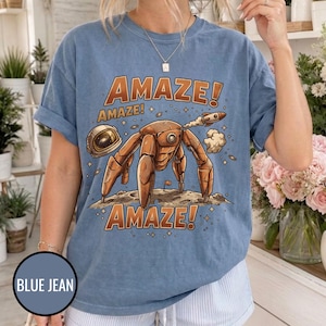May include: A blue short-sleeve t-shirt featuring a graphic of a brown robotic creature on a moon surface with an astronaut helmet and a small rocket. The text AMAZE! is printed in large orange letters above and below the central illustration.