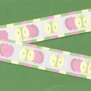 May include: White ribbon with a repeating pattern of pink and yellow apples. The ribbon has green and yellow striped edges. The ribbon is displayed on a green background.