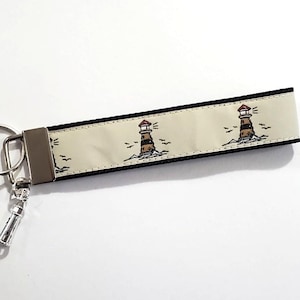 May include: A cream-coloured keychain wristlet with a repeating lighthouse pattern. The lighthouses are brown and red, set against a sea with small birds. The wristlet has a silver-coloured metal clasp and key ring.