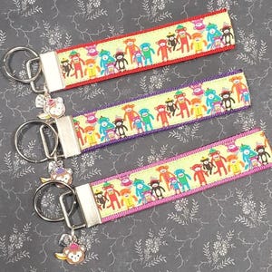 Op de afbeelding: Drie kleurrijke sleutelhangers met aapmotieven. Elke sleutelhanger heeft een ander gekleurd lint met een herhalend patroon van apen die hoeden dragen en bananen vasthouden. De sleutelhangers zijn bevestigd aan zilveren sleutelringen met een aapbedel.