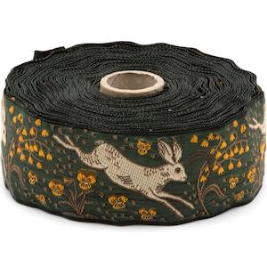 May include: A roll of dark green woven ribbon featuring a repeating pattern of leaping cream-colored hares and small yellow flowers. The ribbon has a textured surface and a central cardboard core, displaying a detailed nature-inspired design.