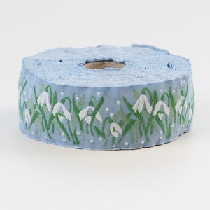 May include: A roll of light blue ribbon with a white and green floral pattern. The ribbon features a repeating design of white snowdrop flowers with green leaves.