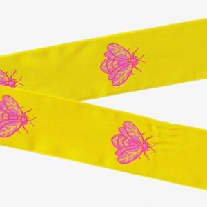 May include: A bright yellow fabric ribbon featuring a repeating pattern of embroidered pink moths with yellow wing details. The ribbon is positioned in a V-shape, showcasing the smooth texture of the material and the consistent insect design.