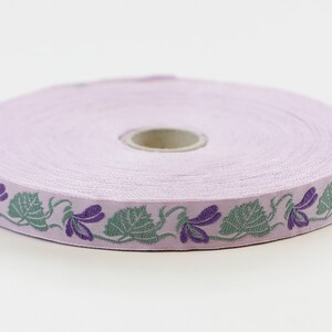 Cinta jacquard KAFKA C-01/04 de algodón orgánico tejido, 16 mm de ancho, fondo lila con violetas moradas y hojas/enredaderas verdes.