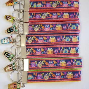May include: A collection of keychains with colorful owl designs. The keychains feature a navy blue strap with an owl pattern in various colors, including orange, yellow, and green. Each keychain has a metal clasp and a decorative owl charm.