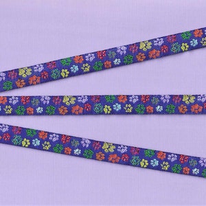 May include: Three purple ribbons with a repeating pattern of colorful paw prints. The ribbons are arranged in a staggered pattern on a purple background.