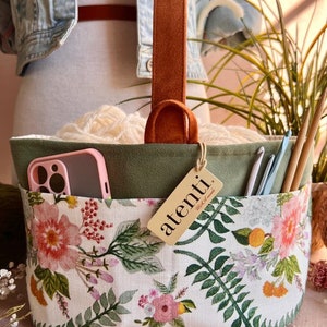 May include: A floral print craft organizer with a brown leather handle. The interior features a green lining and holds white yarn, crochet hooks, and a pink phone case. The tag reads "atenti."