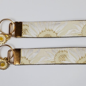 May include: Two keychains with cream-colored fabric straps featuring a sunflower design in white and gold. Each keychain has a gold-colored metal ring, a gold-colored metal clasp, and a sunflower charm.