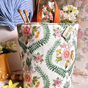 May include: A white tote bag with a floral pattern featuring pink, orange, and yellow flowers, and green leaves. The bag has orange handles and a tag that says "atenti". Two wooden knitting needles are inside the bag.