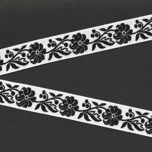 May include: White ribbon with a black floral pattern. The design features stylized flowers, leaves, and berries. The ribbon is angled across a dark background, showcasing the detailed embroidery.