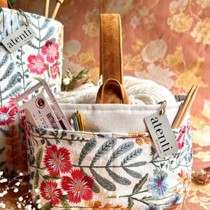 May include: Embroidered fabric knitting basket with a brown leather handle. The basket is filled with white yarn, knitting needles, and other craft supplies. The fabric features red, blue, and green floral embroidery. The tag reads "atenti."