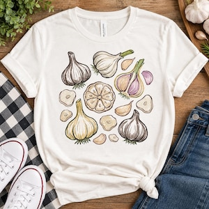 Puede incluir: Una camiseta blanca de manga corta con un estampado gráfico de varios bulbos de ajo, dientes y rodajas en tonos blanco, beige y morado. La ilustración muestra bulbos de ajo enteros, una sección transversal y varios dientes de ajo individuales.