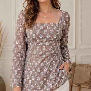 May include: A long-sleeved, taupe-coloured tunic top featuring a square neckline and a repeating white floral pattern. The garment includes side pockets and is paired with white trousers, creating a cohesive look with a structured fit.