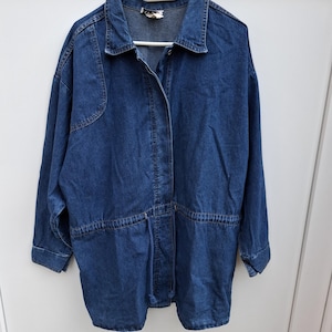 Puede incluir: Una chaqueta de mezclilla azul oscuro con cuello clásico, mangas largas con puños abotonados y una cintura con cordón ajustable en azul a juego. La chaqueta presenta un cierre frontal de botones y un corte estructurado y holgado.