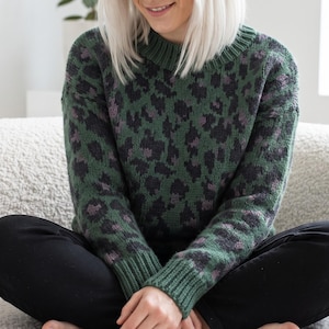 Peut inclure: Un pull en tricot à manches longues présentant une base vert foncé avec un motif léopard noir et violet sourd. Le vêtement possède un col rond côtelé et des poignets côtelés au bout des manches, offrant une coupe structurée au cou et aux poignets.
