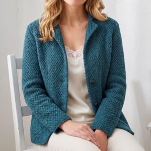 May include: A teal blazer featuring a textured diamond-patterned knit fabric, a notched lapel, and two dark buttons on the front. The blazer is worn over a cream-coloured camisole with lace trim and paired with light-coloured trousers.