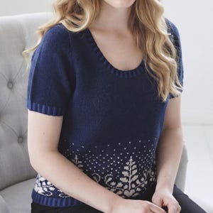 May include: A short-sleeved navy blue knit top featuring a scoop neckline with ribbed trim. The lower section of the garment displays a white patterned design of stylised trees and foliage, finished with a matching ribbed hem at the bottom.