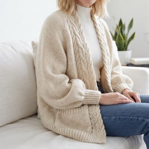Peut inclure: Un gilet en tricot épais de couleur crème avec un motif torsadé le long de l'ouverture avant, des manches longues à poignets côtelés et deux poches avant. Le gilet est porté sur un haut blanc à col montant côtelé et un jean bleu.
