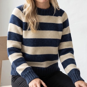 May include: A long-sleeved knit jumper featuring horizontal stripes in navy blue and cream. The garment has a ribbed crew neckline, ribbed cuffs at the wrists, and a ribbed hemline. The knit texture is visible throughout the entire jumper.