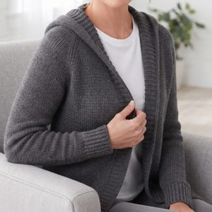 May include: A dark charcoal grey knit hooded cardigan featuring a textured stitch pattern, long sleeves with ribbed cuffs, and a wide ribbed front border. The garment is worn over a plain white t-shirt, showcasing a casual and open-front design.