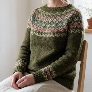Peut inclure: Un pull en tricot vert olive à manches longues avec un col rond et des poignets côtelés. Le vêtement présente un empiècement détaillé et des poignets décorés d'un motif floral et géométrique multicolore dans des tons rose, crème et bleu.