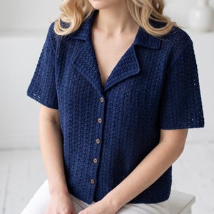 May include: A dark navy blue short-sleeved shirt featuring an open collar and a front button-down closure. The garment is crafted with a textured, open-work crochet knit pattern throughout, showcasing a repeating geometric design in the fabric.