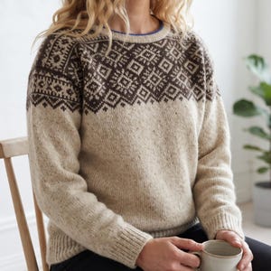 Pode incluir: Uma camisola de malha de manga comprida com uma base de cor creme e um padrão geométrico castanho escuro no jugo e nos ombros. A camisola tem um decote redondo com um rebordo interior azul e punhos canelados nos pulsos e na bainha.