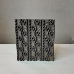 May include: A rectangular dark brown wooden block for textile printing featuring a repeating pattern of vertical wavy lines and rows of small diamond shapes. The surface has a textured, weathered appearance with carved geometric details.