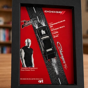 Könnte beinhalten: Ein gerahmtes Display mit einem schwarzen Dodge Charger R/T Modellauto auf einer rot-schwarzen Straßengrafik. Es enthält Text für The Fast and the Furious, Dodge, Charger R/T, Motorspezifikationen und ein Bild von Dominic Toretto.
