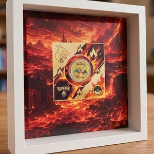 Könnte beinhalten: Ein weißer Schattenfugenrahmen mit einer quadratischen Leinwand, die eine feurige Vulkanlandschaft zeigt. Eine runde goldfarbene Münze mit dem Charakter Glurak ist mittig auf der Leinwand platziert, umgeben von Flammensymbolen, dem Text Pokemon und der Zahl 6.