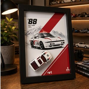 Può includere: Un'opera d'arte da parete incorniciata in 3D che presenta un'illustrazione di un'auto da rally Mitsubishi Starion bianca con il testo '88 RALLY LEGEND GROUP A' e un modellino di auto bianco abbinato montato sulla superficie su uno sfondo grafico rosso e bianco.