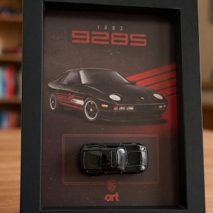 Può includere: Una cornice nera con una stampa di una Porsche 928S del 1983 e un'auto giocattolo nera abbinata montata al centro. Lo sfondo è rosso scuro e nero con il testo 1983 928S e un piccolo logo art in basso. L'auto è mostrata con una vista dall'alto.