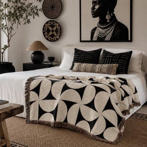 May include: A woven throw blanket featuring a repeating black and cream geometric pattern of interlocking semi-circles and fan shapes, finished with a short fringe along the edges, draped over a white bedspread in a bedroom setting.