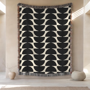 May include: A woven throw blanket featuring a repeating geometric pattern of black semicircles on a cream background. The blanket has a textured surface and is finished with a decorative fringe along the top and bottom edges.