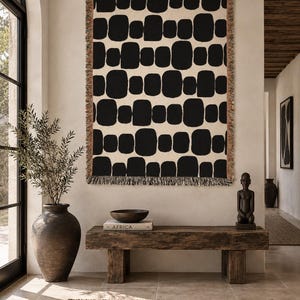 May include: A rectangular woven throw blanket featuring a repeating pattern of rounded black shapes on a cream background. The blanket has a fringed edge with multi-colored threads and is displayed hanging vertically on a wall as a decorative piece.