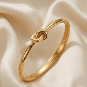 May include: A polished gold-tone bangle bracelet featuring a central knot design. The circular band has a smooth, reflective surface and a hinged closure mechanism, showcasing a continuous, sleek metallic finish throughout the entire piece.