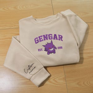以下が含まれることがあります： ベージュのクルーネックトレーナー。紫色の刺繍でキャラクターのゲンガーと「GENGAR EST. 1996」の文字が施されています。袖口には黒い筆記体で「Custom name」という文字が刺繍されています。