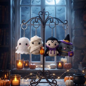 Puede incluir: Cuatro llaveros de Halloween tejidos a ganchillo colgados de un soporte de metal oscuro. Las figuras incluyen un fantasma blanco, una momia beige con una calabaza naranja, un vampiro con capa negra y lazo morado, y un gato gris con sombrero.