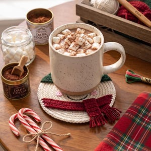 May include: A speckled ceramic mug filled with hot cocoa and marshmallows sits on a crocheted snowman coaster. Nearby are two candy canes tied with twine, a jar of marshmallows, two tins of dark cocoa, a wooden crate with yarn, and a plaid napkin.