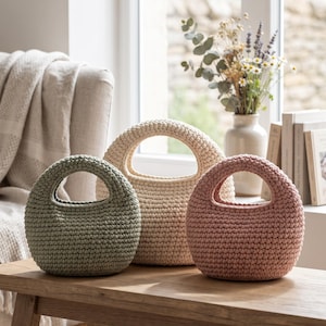 May include: Three round, crocheted handbags with integrated circular handles. The bags are arranged in a row, featuring a sage green bag on the left, a cream-colored bag in the center, and a dusty rose pink bag on the right, all with a textured stitch.