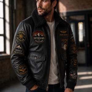 Puede incluir: Una chaqueta bomber de cuero negro con cuello de piel sintética oscura, puños acanalados y múltiples parches bordados que incluyen textos como Skybound Expeditions, Aero Legacy, Freedom Flight, True North y Higher Than Yesterday.