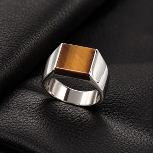 May include: A polished silver ring featuring a square, flat-topped tiger's eye gemstone inlay. The band is smooth and metallic, showcasing a reflective finish that contrasts with the warm, golden-brown tones of the natural stone centerpiece.