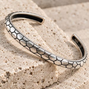 May include: A silver cuff bracelet featuring a repeating hexagonal pattern across the outer surface. The metal has a polished finish with darkened recessed lines between the shapes. The interior of the band is stamped with the number 925.
