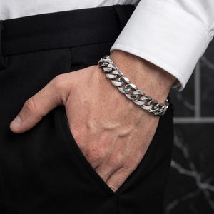 Könnte beinhalten: Ein poliertes Panzerkettenarmband aus silberfarbenem Metall mit dicken, ineinandergreifenden flachen Gliedern. Das Armband hat eine glänzende metallische Oberfläche und einen sicheren Verschluss, getragen am Handgelenk zu weißer Manschette und schwarzer Hose.